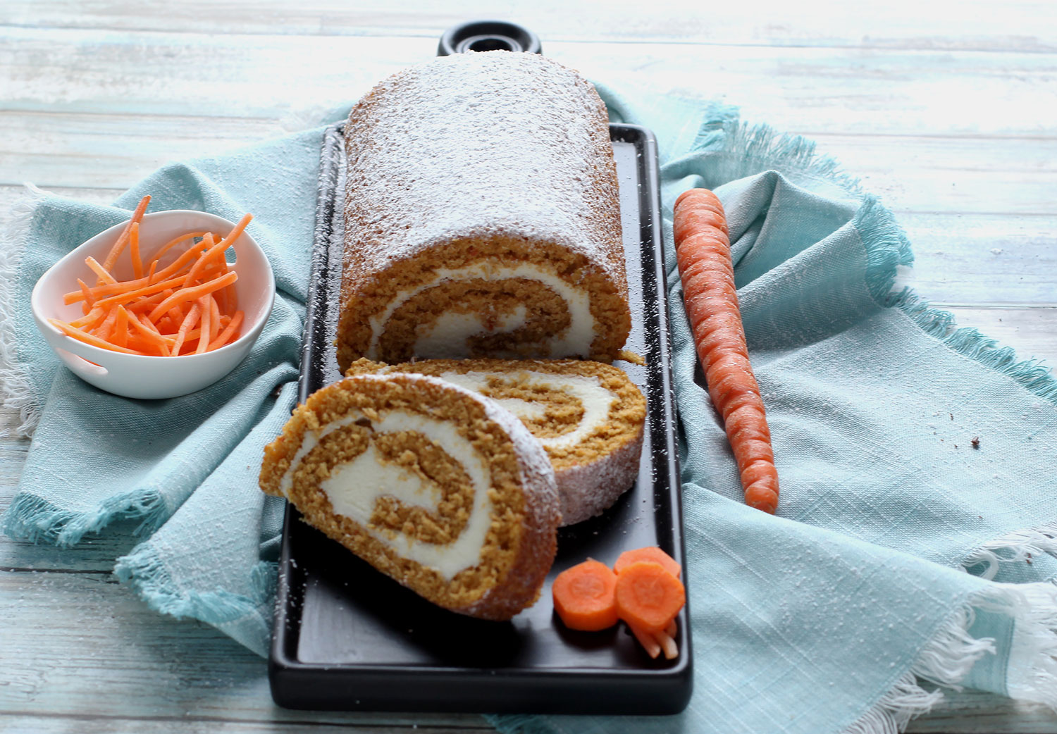 Carrot Cake Roll and slices on cutting board next to whole carrot and bowl of carrot shavings
