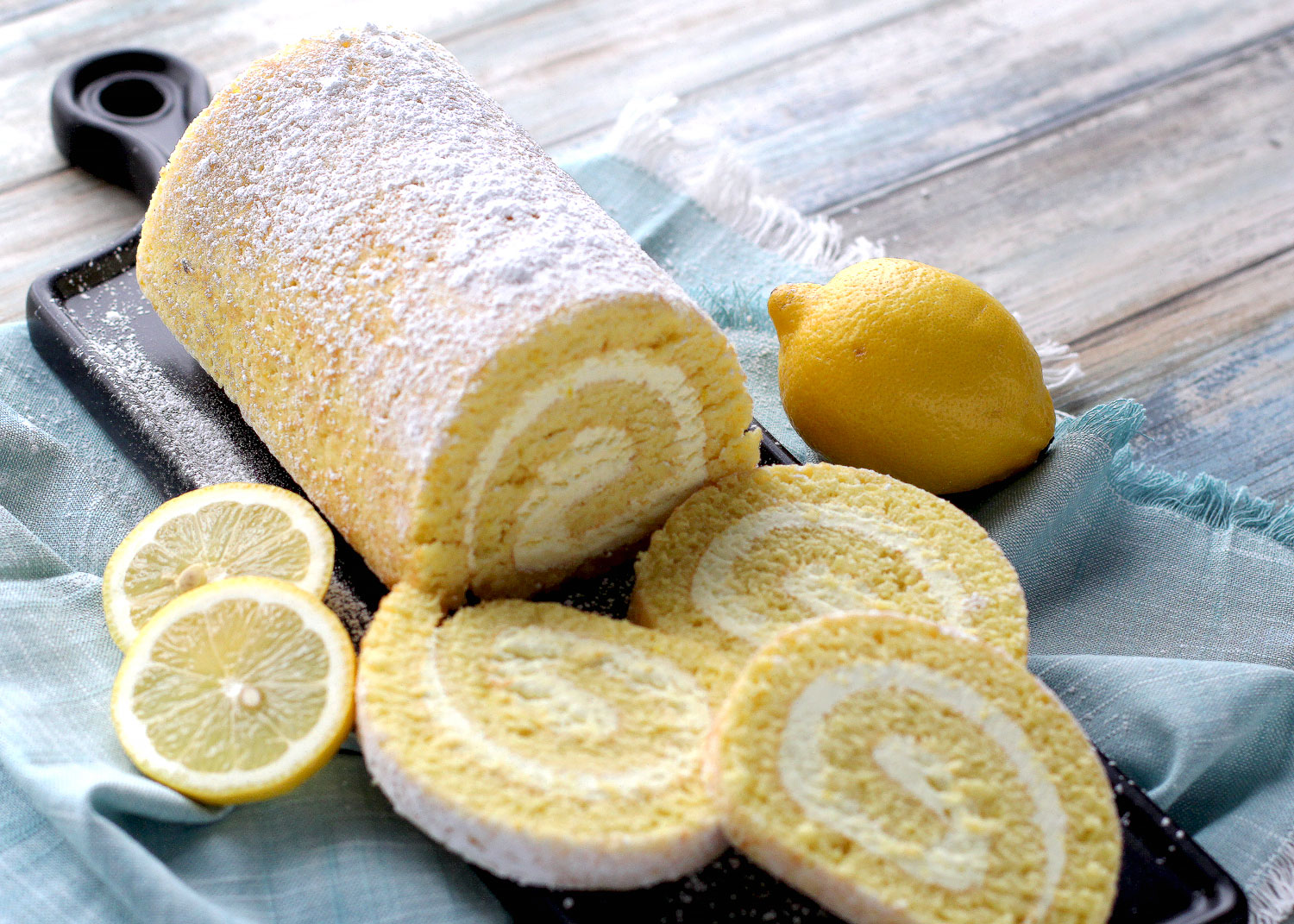 Lemon Crème cake roll and slices on cutting board next to a lemon and lemon slices