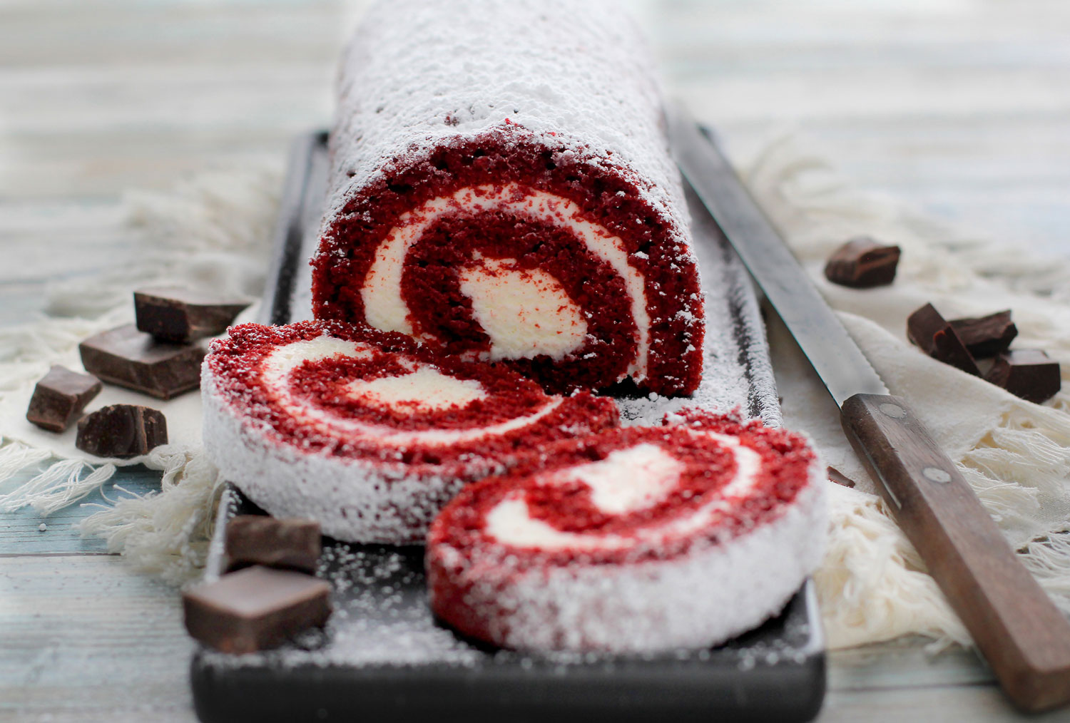 Red velvet cake roll slices on cutting board with knife and chocolate pieces