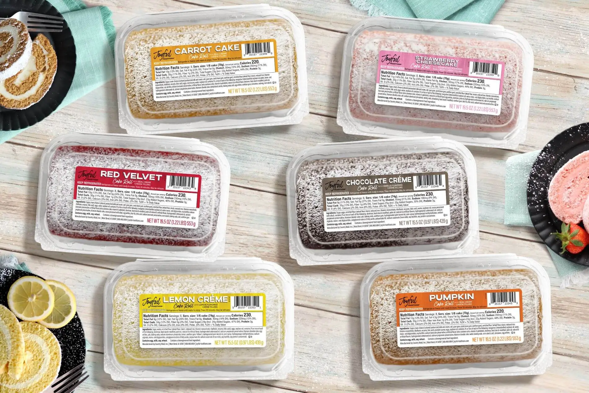 All flavors of Joyful Traditions Cake Rolls in clamshells with labels on wooden background with plates of cake rolls next to them
