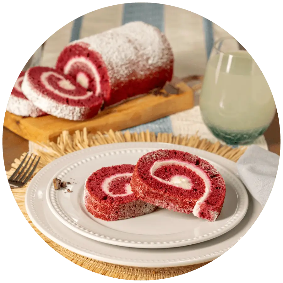 Joyful Traditions Red Velvet Cake Roll - two slices on a plate with the full cake roll on a cutting board in the background.