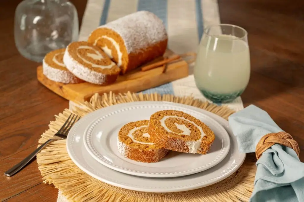 Joyful Traditions Pumpkin Cake Roll - two slices on a plate with the full cake roll on a cutting board in the background.