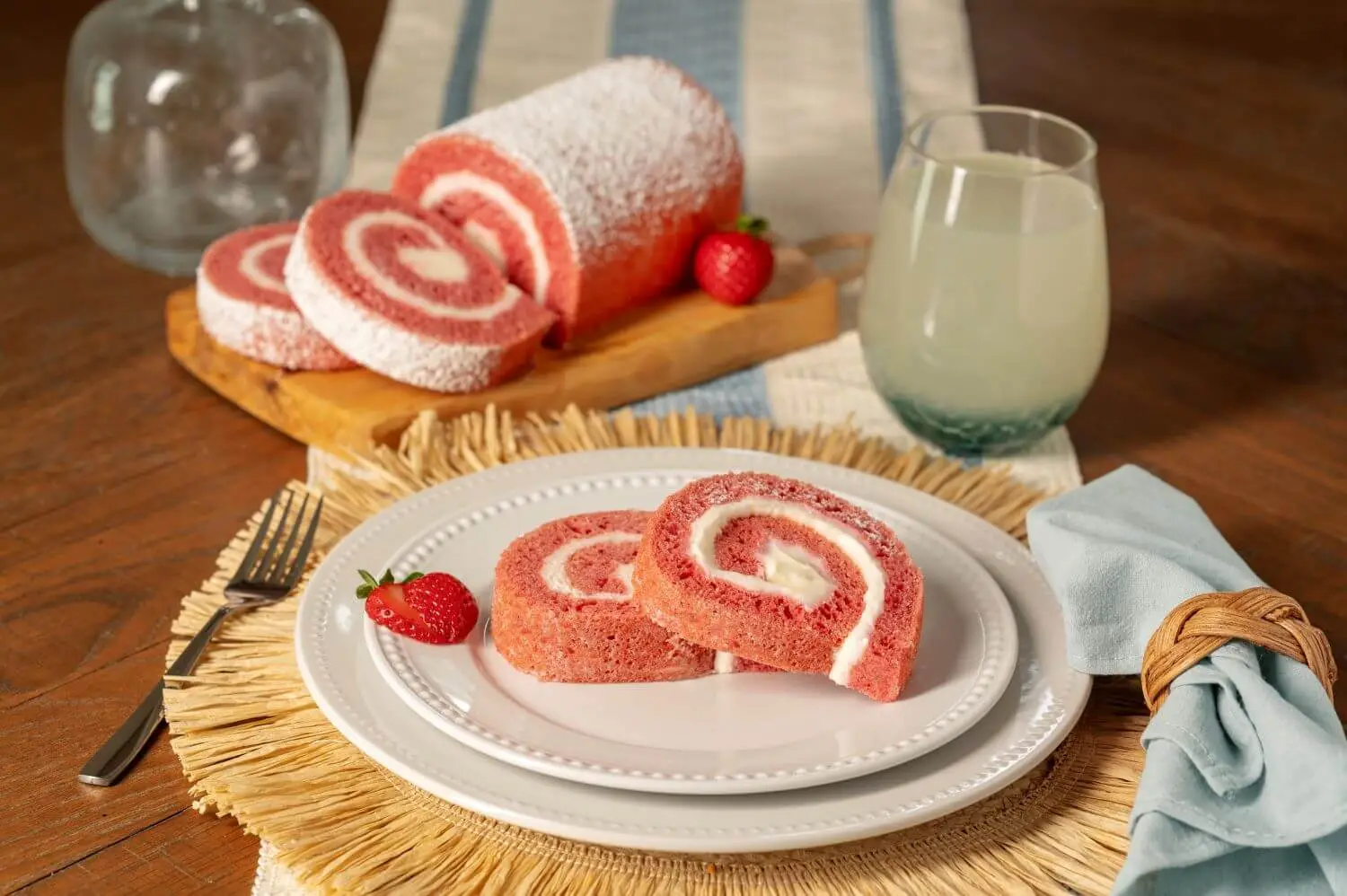 Joyful Traditions Strawberry Cheesecake Cake Roll - two slices on a plate with the full cake roll on a cutting board in the background.